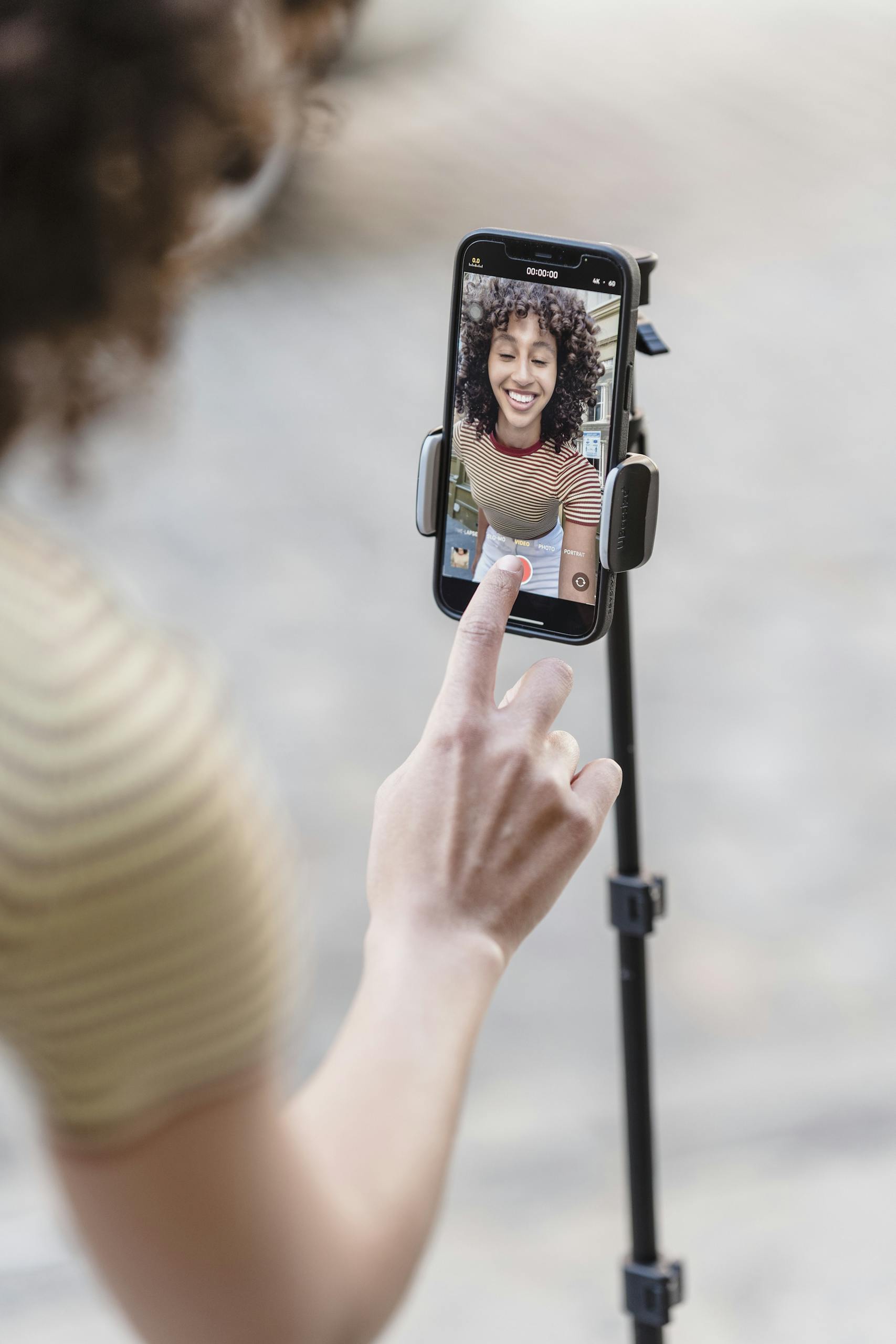 Crop positive ethnic female touching screen of smartphone while recording live video on street with paved sidewalk on blurred background