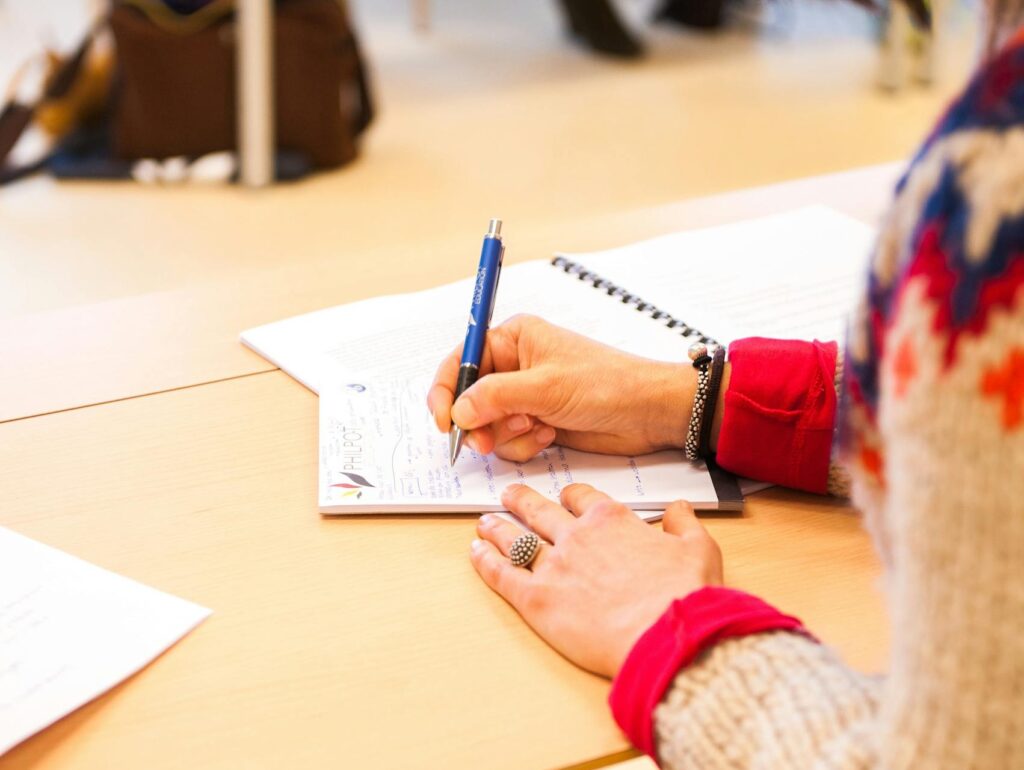 A woman writing notes in a notebook during a classroom lecture setting.